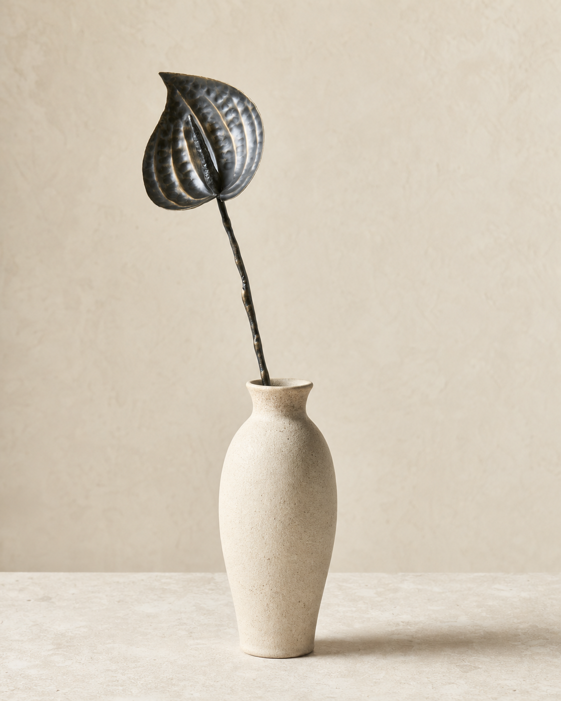 Single aged brass anthurium stem displayed in a neutral ceramic vessel.
